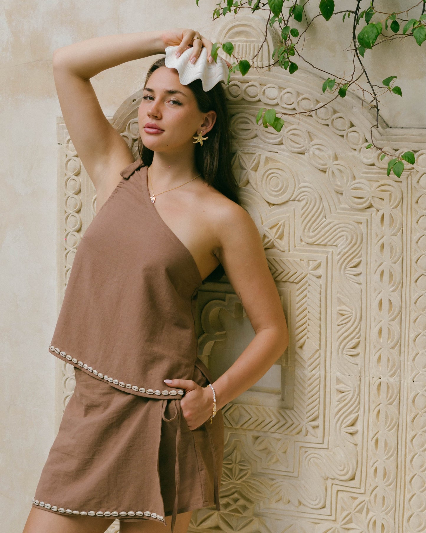 The Coralie Shell Top in chocolate brown features a statement tie-up shoulder, side slits, and delicate cowrie shell trim for a breezy coastal look.