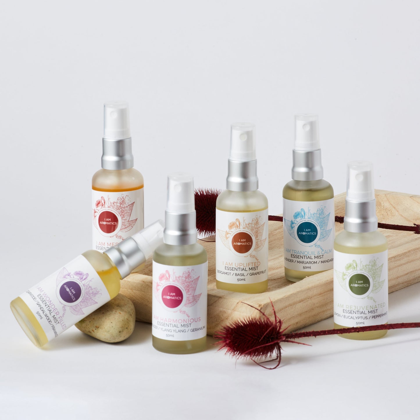 Essential Mist collection, 6 bottles with coloured botanical logos, frosted white glass, matt silver atomiser, leaning on a wooden try with a rock and 2 dryed flower branches