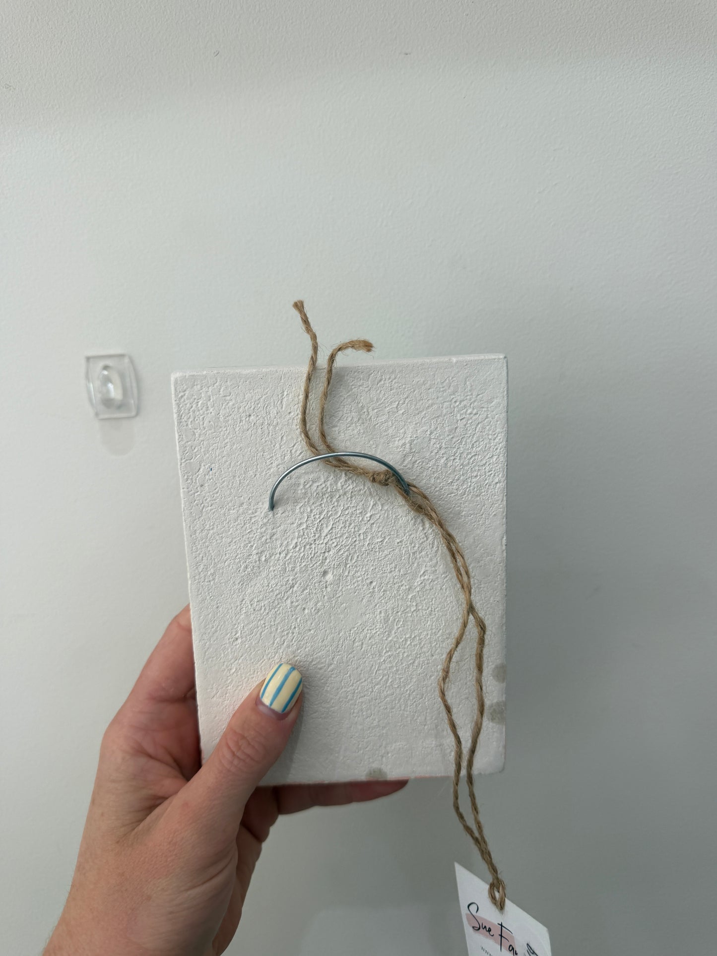 A hand holding a square white textured box or plaque with a rustic brown twine loop and a small hanging tag, photographed against a plain light gray wall.